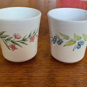 Pode incluir: Duas canecas de cerâmica branca com desenhos florais. Uma caneca tem flores cor-de-rosa e folhas verdes, enquanto a outra tem flores azuis e folhas verdes.