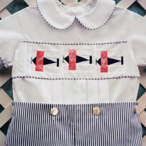 May include: A white short-sleeved shirt with a rounded collar and a smocked chest panel featuring three embroidered black and red airplane motifs. The lower section consists of navy blue and white vertical pinstriped fabric with two buttons.