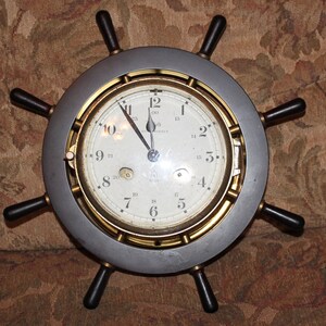 May include: A ship's wheel clock with a silver-colored frame and a gold-colored inner ring. The clock face is white with black numerals and hands. The wheel's handles are dark brown.