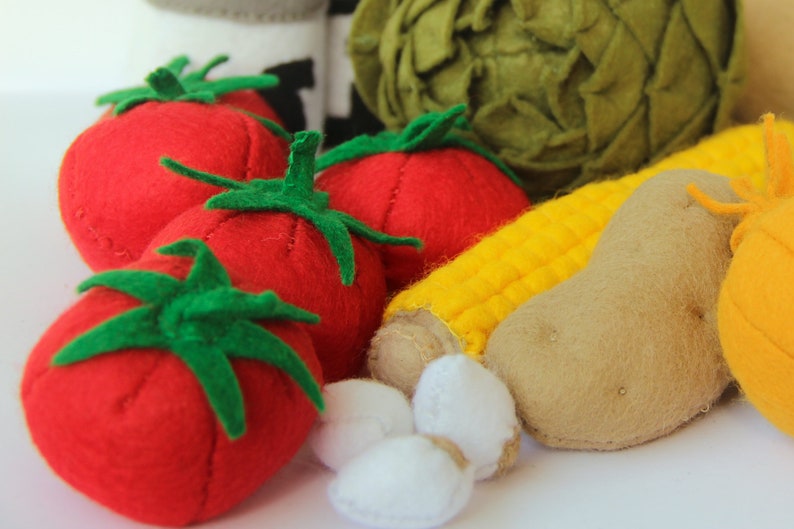 Pu&ograve; includere: Una collezione di alimenti giocattolo in feltro che include tre pomodori rosso brillante con piccioli verdi, una pannocchia di mais gialla, una patata color marrone chiaro, un carciofo verde rotondo e piccoli pezzi bianchi, realizzati in tessuto.