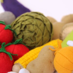 Pu&ograve; includere: Una collezione di verdure in feltro che include tre pomodori rossi con gambi verdi, un carciofo verde scuro, una pannocchia di mais gialla, una patata marrone, una cipolla arancione, una melanzana viola e un gambo di sedano verde chiaro.