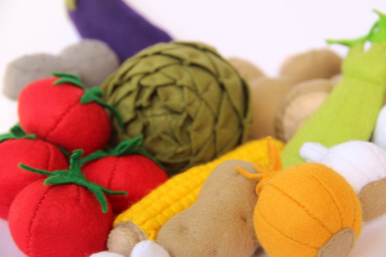 Pu&ograve; includere: Una collezione di verdure in feltro che include tre pomodori rossi con gambi verdi, un carciofo verde scuro, una pannocchia di mais gialla, una patata marrone, una cipolla arancione, una melanzana viola e un gambo di sedano verde chiaro.