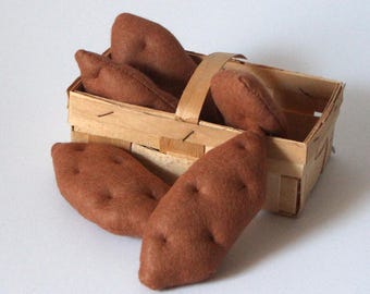May include: A rectangular wooden basket filled with several brown, plush, potato-shaped items. The fabric potatoes feature small, stitched indentations on their surfaces, mimicking the appearance of natural tubers, and are arranged both inside and outside the basket.