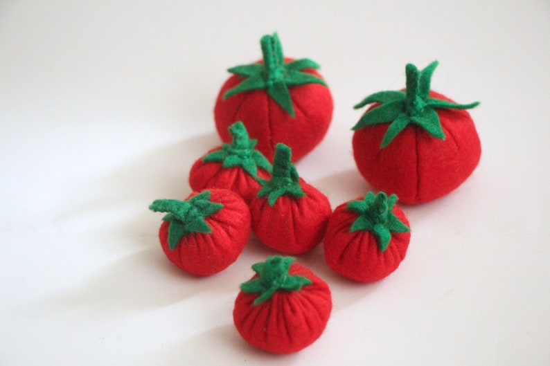 Pu&ograve; includere: Una collezione di sei piccoli pomodori rotondi in feltro rosso, ognuno dotato di un gambo e foglie verdi in feltro. I pomodori variano leggermente nelle dimensioni e sono disposti in un gruppo, mostrando una morbida struttura in tessuto.