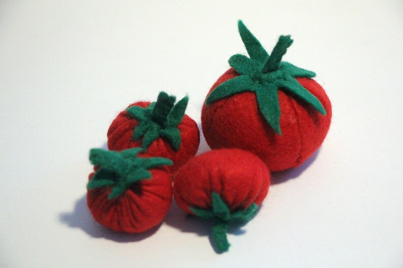 Pu&ograve; includere: Una collezione di quattro pomodori in feltro fatti a mano di varie dimensioni. Ogni pomodoro rosso rotondo presenta un gambo e una struttura a foglie in feltro verde sulla parte superiore. I morbidi giocattoli in tessuto sono raggruppati.