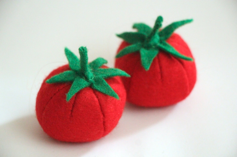 Pu&ograve; includere: Due puntaspilli rotondi in feltro rosso a forma di pomodoro. Ogni puntaspillo presenta un gambo e delle foglie in feltro verde fissati al centro. Il tessuto ha un aspetto morbido e strutturato con segmenti cuciti che creano una forma rotonda.