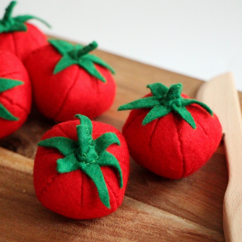 Pu&ograve; includere: Una collezione di quattro pomodori in feltro realizzati a mano, ognuno caratterizzato da un corpo arrotondato rosso vivace e da un distinto picciolo verde a forma di stella sulla parte superiore. Le verdure in tessuto morbido mostrano la trama.
