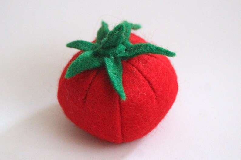 Pu&ograve; includere: Un puntaspilli in feltro rosso a forma di pomodoro, caratterizzato da una superficie testurizzata e da un picciolo in feltro verde con sei foglie appuntite sulla parte superiore. Il corpo presenta sottili rientranze verticali.