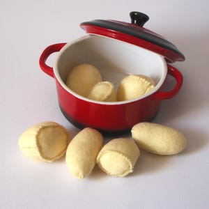 May include: A small red and black ceramic pot with a lid, filled with several pale yellow felt dumplings. Several more dumplings are scattered around the pot, creating a playful, whimsical scene.