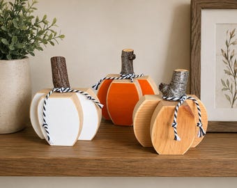 May include: Three decorative wooden pumpkins with natural bark stems and black and white twine bows. One pumpkin is painted white, one is painted bright orange, and one features a natural wood grain finish, all arranged on a flat surface.