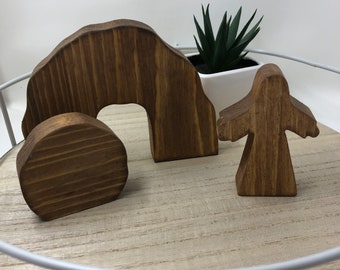 May include: Wooden Easter set with a brown tomb, a round stone, and a standing figure. The tomb is shaped like an archway and the figure is standing with arms outstretched.