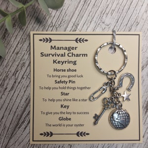 May include: A silver keyring with a horseshoe, safety pin, star, key, and globe charm. The card attached to the keyring reads "Manager Survival Charm Keyring" and lists the meaning of each charm.