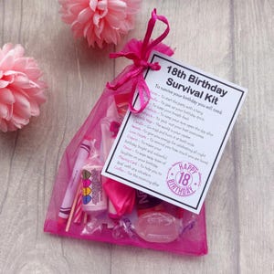 May include: A pink mesh gift bag tied with a ribbon, containing various small items. A card reads "18th Birthday Survival Kit" with a list of items like a balloon, sweets, and a crayon. Pink paper flowers are in the background.