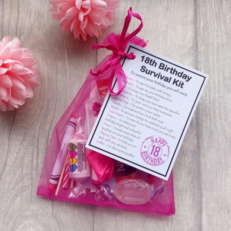 May include: A pink mesh gift bag tied with a ribbon, containing various small items. A card reads "18th Birthday Survival Kit" with a list of items like a balloon, sweets, and a crayon. Pink paper flowers are in the background.