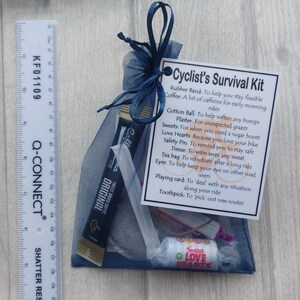 May include: A navy blue mesh bag tied with a dark blue ribbon, containing a cyclist's survival kit. The kit includes coffee, sweets, a safety pin, and a playing card. A ruler is placed to the left of the bag.