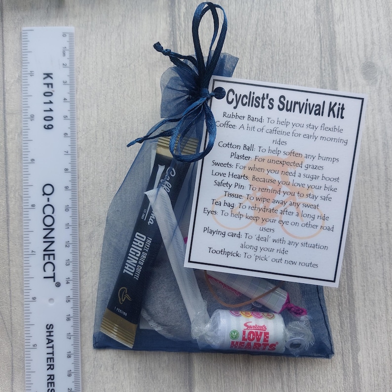 May include: A navy blue mesh bag tied with a dark blue ribbon, containing a cyclist's survival kit. The kit includes coffee, sweets, a safety pin, and a playing card. A ruler is placed to the left of the bag.