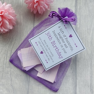 May include: A purple organza bag filled with small pink paper slips, each containing a quote. The bag has a white card attached with the text "A Gift of Positivity, Laughter and Inspiration on your 30th Birthday. Every day for a month, pick a quote to help you reflect on life and brighten each day."