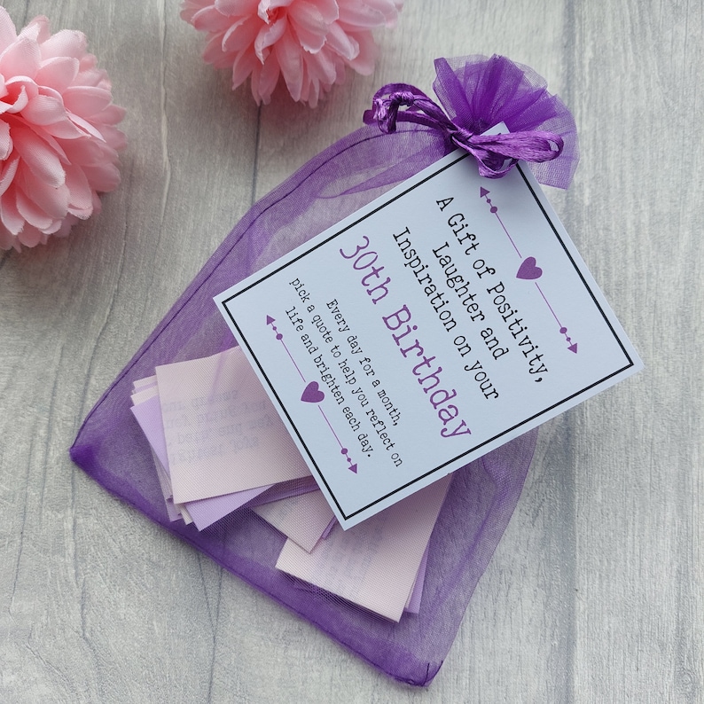 May include: A purple organza bag filled with small pink paper slips, each containing a quote. The bag has a white card attached with the text "A Gift of Positivity, Laughter and Inspiration on your 30th Birthday. Every day for a month, pick a quote to help you reflect on life and brighten each day."