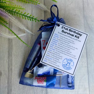 May include: A blue organza bag filled with a 21st birthday survival kit. The kit includes a balloon, umbrella, mint, matchsticks, toothpick, world map, candle, sweets, love hearts, crayon, tissue, playing card, and coffee. The bag has a tag that reads "21st Birthday Survival Kit" and lists the items included with their uses.