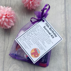 May include: A purple organza bag filled with a 70th birthday survival kit. The kit includes a candle, balloon, cotton wool, crayon, world map, paperclip, cocktail umbrella, sweets, love hearts, coffee, playing card, tea bag, mint, jigsaw piece, and tissue. The bag has a tag attached that reads "70th Birthday Survival Kit" and lists the contents of the kit.