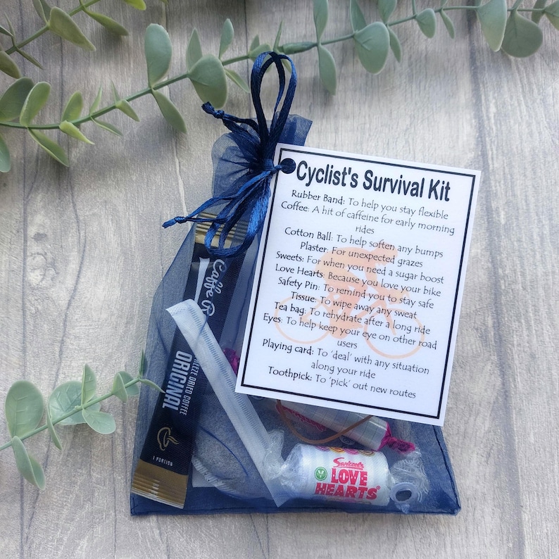 May include: A navy blue mesh gift bag tied with a matching ribbon, containing a cyclist's survival kit. The kit includes coffee, sweets, a safety pin, and a card with text. The bag is presented on a light wooden surface with green foliage.