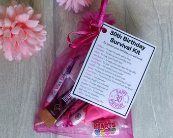 May include: A pink organza bag filled with a 30th birthday survival kit. The kit includes a balloon, umbrella, mint, matchstick, toothpick, world map, candle, sweets, love hearts, crayon, tissue, playing card, and coffee. The bag also includes a card with the text "30th Birthday Survival Kit" and a list of items needed to survive the birthday.