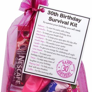 May include: A pink tulle bag filled with a birthday survival kit. The kit includes a balloon, umbrella, mint, matchsticks, toothpick, world map, candle, sweets, love hearts, crayon, tissue, playing card, and coffee. The bag has a tag attached that reads "30th Birthday Survival Kit" and lists the items included in the kit with their uses. The tag also has a Happy 30th Birthday stamp.