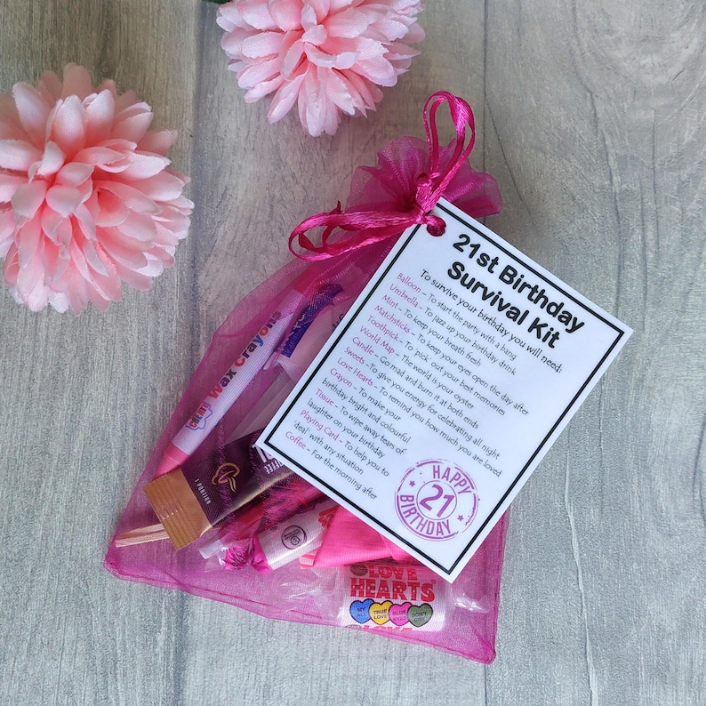 May include: A pink and white gift bag filled with a 21st birthday survival kit. The kit includes a card with a list of items and their uses, such as balloons, matches, and sweets. The card also has the text "Happy 21st Birthday".