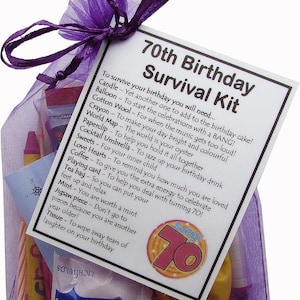 May include: A purple organza bag filled with a 70th birthday survival kit. The kit includes a candle, balloon, cotton wool, crayon, world map, paperclip, cocktail umbrella, sweets, love hearts, coffee, playing card, tea bag, mint, jigsaw piece, and tissue. The bag has a white card attached with the text "70th Birthday Survival Kit" and a list of items needed to survive the birthday.