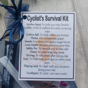 May include: A Cyclist's Survival Kit gift set in a sheer blue bag, tied with a blue ribbon. The kit includes a card with a list of items like a rubber band, coffee, cotton ball, plaster, sweets, and a playing card.