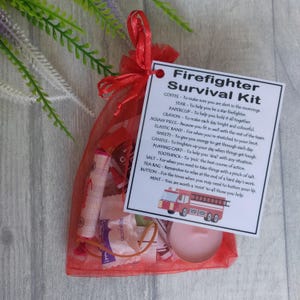 May include: A red mesh bag filled with a firefighter survival kit. The kit includes a small candle, a tea bag, a paperclip, a star, a crayon, a toothpick, a button, a rubber band, a small bag of sweets, and a small bag of salt. The bag is tied with a red ribbon. The kit comes with a card that lists the items and their uses. The card reads "Firefighter Survival Kit".
