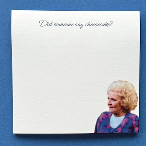 May include: A white notepad with the text "Did someone say cheesecake?" in a script font. The notepad features a color image of a woman with blonde hair and a blue and purple patterned top. The notepad is on a blue surface.