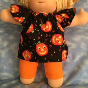 May include: A soft doll with blonde yarn hair wearing a black short-sleeved top featuring a repeating orange jack-o'-lantern pumpkin pattern with small green dots and red swirls, paired with solid orange leggings that reach the mid-thigh area.