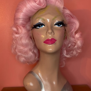 May include: A mannequin head with a pink wig styled in voluminous curls. The mannequin has blue eyes, long eyelashes, and bright pink lipstick. The background is a solid coral color.