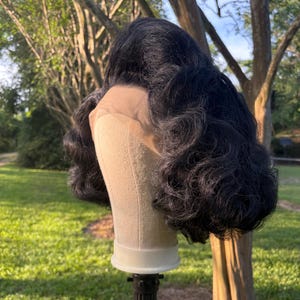May include: A black wig with voluminous, curly hair is displayed on a mannequin head. The wig features a lace front and is set against a backdrop of green grass and trees, suggesting an outdoor setting.