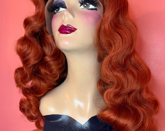 May include: A mannequin head with a vibrant, wavy, auburn wig. The mannequin has painted features, including dark eyebrows, long eyelashes, and bright pink blush. The wig is styled with voluminous curls.