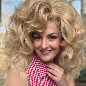 May include: A woman with a large, voluminous blonde wig and a red and white checkered shirt smiles at the camera. The wig has large, soft curls. The shirt is a classic gingham pattern.