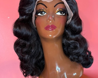 May include: A black wig with voluminous, wavy curls styled on a mannequin head. The wig features a lace front and is set against a pink background. The mannequin has painted features, including gold eyeshadow and red lipstick.