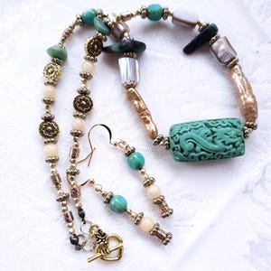 May include: A necklace and earring set featuring a variety of beads in shades of gold, cream, turquoise, and green. The necklace has a large, carved turquoise rectangular centerpiece. The earrings have turquoise and cream-colored beads.