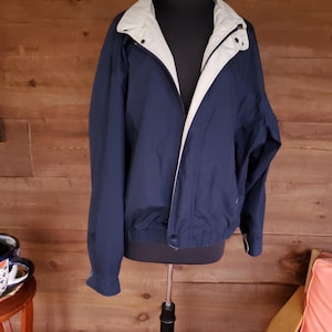 May include: A navy blue jacket with a light beige collar and lining, displayed on a black mannequin. The jacket has a zipper closure and a button at the waist. The background is a wooden wall.