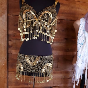 May include: A black and gold belly dance costume displayed on a mannequin. The top and skirt are heavily embellished with gold sequins, beads, and dangling coins. The costume has a decorative floral and geometric pattern.