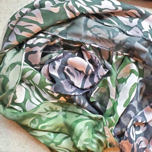 May include: A folded silk scarf with a botanical pattern in shades of green, gray, and pale pink. The scarf has a flowing, layered appearance, with the pattern repeating across the fabric. The scarf appears to be made of a lightweight material.