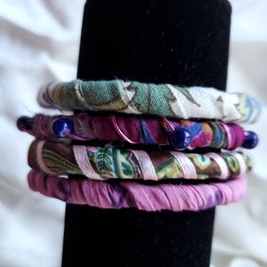 May include: A stack of fabric-covered bangles in various colors and patterns. The bangles are wrapped in floral and paisley fabrics, with accents of pink and blue. Some bangles have blue beads and wire accents. The bangles are displayed on a black stand.