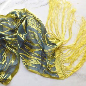 May include: A flowing scarf in shades of teal and yellow. The teal fabric is patterned with gold leaf-like designs. The edges and fringe are a bright yellow. The scarf is made of a lightweight, possibly silk, material.