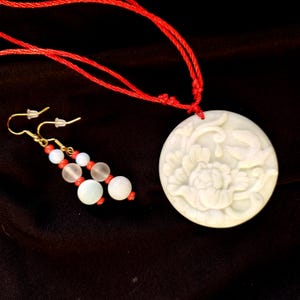 May include: A white pendant necklace with a carved floral design and a red cord. The image also shows a pair of earrings with gold-colored hooks and a combination of white, red, and clear beads. The jewelry is set against a dark background.