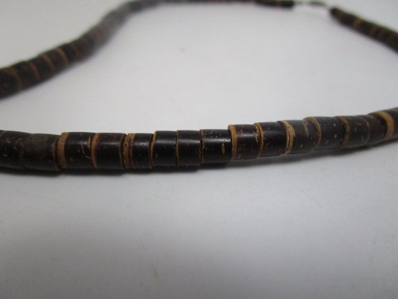 May include: A close-up of a necklace made of dark brown wooden beads. The beads are cylindrical and have a slightly rough texture.