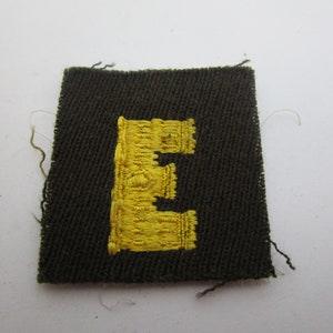 May include: A dark green fabric patch with a yellow embroidered letter "E" in the center.