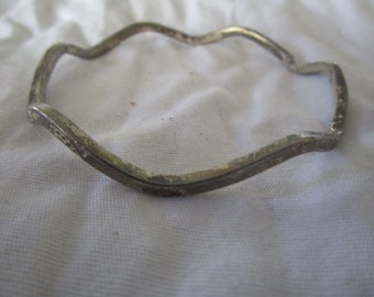 May include: A silver-toned, wavy-edged bracelet. The bracelet has a unique, irregular shape, with a series of curves and angles. The metal appears to have a slightly aged or textured finish, adding to its vintage appeal. The bracelet is resting on a white surface.