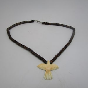 May include: A brown beaded necklace with a carved bone bird pendant. The bird is white with a light brown color on the wings.