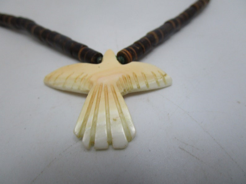 May include: A necklace with a carved bird pendant made of white shell. The necklace is made of brown wooden beads.